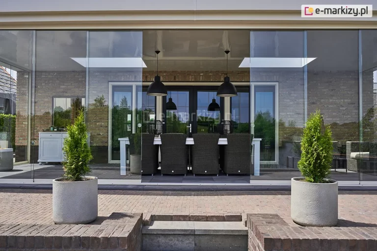 Szeroka zabudowa tarasu z systemem bezramowych szklanych drzwi przesuwnych Auris, zamontowana przy budynku z jasnej, tradycyjnej cegły. Zdjęcie prezentuje frontowy widok na przeszkloną jadalnię letnią. Centralna część szklanej ściany jest rozsunięta, ukazując wnętrze z dużym białym stołem, ciemnymi plecionymi fotelami oraz dwiema czarnymi lampami wiszącymi w stylu industrialnym. Przeszklona konstrukcja posiada dach zintegrowany z elewacją, a na szybach widoczne są okrągłe, metalowe uchwyty do przesuwania paneli. Przed zabudową znajduje się taras z czerwonej kostki brukowej, na którym ustawiono dwie nowoczesne, betonowe donice z zielonymi iglakami, flankujące wejście.