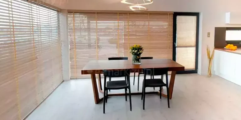 Zdjęcie prezentuje jasną i przestronną jadalnię połączoną z kuchnią, w której głównym elementem osłonowym są żaluzje drewniane 25 mm zamontowane na dużych przeszkleniach narożnych. Żaluzje mają naturalny, jasny odcień drewna i są wyposażone w taśmowe drabinki w kolorze zbliżonym do lameli, które przebiegają pionowo przez całą wysokość osłon. Dzięki poziomemu ustawieniu listewek do wnętrza wpada rozproszone światło dzienne, tworząc przytulną atmosferę. Na środku pomieszczenia stoi masywny stół jadalniany wykonany z ciemnego drewna o wyraźnym usłojeniu, przy którym ustawiono cztery nowoczesne, czarne krzesła o minimalistycznej formie. Na blacie stołu znajduje się szklany wazon z bukietem żółtych tulipanów. W tle po prawej stronie widoczny jest fragment białej zabudowy kuchennej z ciemnym blatem, na którym leżą pomarańcze, oraz wysokie okno tarasowe z ciemną ramą, również przesłonięte pasującą żaluzją. Podłoga w całym pomieszczeniu jest wyłożona jasnymi panelami o chłodnym odcieniu, co kontrastuje z ciepłą barwą drewna stołu i osłon okiennych. Całość aranżacji dopełnia designerska, biała lampa sufitowa o obłych kształtach zawieszona bezpośrednio nad stołem.