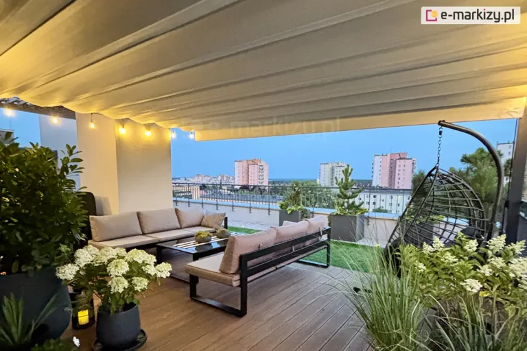 szeroki kadr tarasu widokowego, nad którym zamontowano wielkogabarytowy dach rzymski Supro w kolorze jasnokremowym. Tkanina zadaszenia jest w pełni rozłożona, tworząc jednolity sufit z delikatnymi, poziomymi przetłoczeniami, a pod jej krawędzią zawieszono girlandę świetlną z żarówkami o ciepłej barwie. Przestrzeń wypoczynkowa na tarasie została wyposażona w dwie długie, szare sofy o nowoczesnej, czarnej konstrukcji, ustawione prostopadle do siebie wokół niskiego stolika kawowego ze szklanym blatem. Po prawej stronie widoczny jest czarny fotel wiszący typu kokon, umieszczony na tle metalowej balustrady. Posadzka tarasu składa się z ciemnobrązowych desek kompozytowych oraz fragmentów wyłożonych sztuczną trawą i jasnym żwirem. Aranżację dopełniają liczne rośliny w donicach, w tym kwitnące białe hortensje i ozdobne trawy. W tle rozpościera się szeroka panorama miasta z widocznymi blokami mieszkalnymi pod jasnoniebieskim, wieczornym niebem.