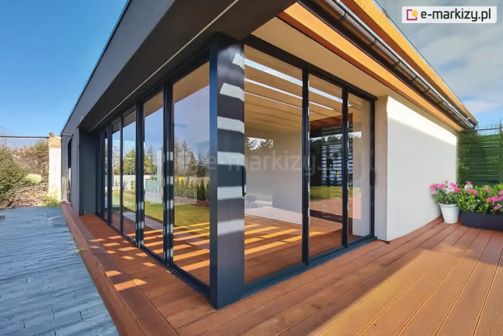 Zdjęcie przedstawia narożny widok na nowoczesną zabudowę tarasu z wykorzystaniem systemu Sotro w kolorze antracytowym. Przeszklone ściany przesuwne łączą się pod kątem prostym, tworząc transparentną osłonę dla przestrzeni wypoczynkowej. Pod konstrukcją dachu widoczne są drewniane lamele w jasnym kolorze, które kontrastują z ciemnymi profilami aluminiowymi. Całość zamontowana jest na szerokim tarasie z desek kompozytowych w kolorze ciepłego brązu. Na tarasie, przy białej ścianie budynku, ustawiono białą donicę z kwitnącymi pelargoniami w kolorze różowym. W szybach odbija się błękitne niebo oraz przydomowa zieleń, co podkreśla efekt przenikania się wnętrza z ogrodem.