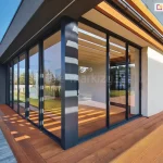 Zdjęcie przedstawia narożny widok na nowoczesną zabudowę tarasu z wykorzystaniem systemu Sotro w kolorze antracytowym. Przeszklone ściany przesuwne łączą się pod kątem prostym, tworząc transparentną osłonę dla przestrzeni wypoczynkowej. Pod konstrukcją dachu widoczne są drewniane lamele w jasnym kolorze, które kontrastują z ciemnymi profilami aluminiowymi. Całość zamontowana jest na szerokim tarasie z desek kompozytowych w kolorze ciepłego brązu. Na tarasie, przy białej ścianie budynku, ustawiono białą donicę z kwitnącymi pelargoniami w kolorze różowym. W szybach odbija się błękitne niebo oraz przydomowa zieleń, co podkreśla efekt przenikania się wnętrza z ogrodem.