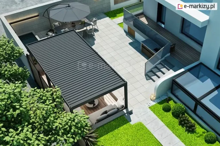 Zdjęcie przedstawia widok z lotu ptaka na nowoczesną aranżację tarasu, w której centralnym punktem jest duża, dwumodułowa pergola wolnostojąca Marlo w kolorze antracytowym. Konstrukcja posiada zadaszenie lamelowe w pozycji całkowicie zamkniętej. Pod pergolą, na drewnianym podeście z brązowych desek, znajduje się strefa wypoczynkowa z szarą kanapą i niskim stolikiem. Taras wyłożony jest jasnoszarymi płytami wielkoformatowymi, a w jego dalszej części umiejscowiono jadalnię pod dużym, szarym parasolem ogrodowym. Całość sąsiaduje z nowoczesnym budynkiem o białej elewacji, wyposażonym w zewnętrzne schody i szklane balustrady. Wzdłuż tarasu rozciąga się pas zieleni z trawnikiem i formowanymi krzewami ozdobnymi, a po lewej stronie kompozycję dopełniają korony drzew liściastych.