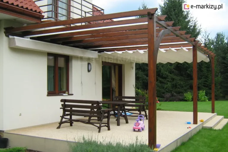 duży, podwyższony taras przydomowy z dwiema niezależnymi sekcjami poziomego dachu rzymskiego Supro, zamontowanymi na wolnostojącej konstrukcji drewnianej. Pergola wykonana jest z belek o ciemnobrązowym wybarwieniu, z ozdobnym łukowym zastrzałem przy jednym z filarów. Pierwsza sekcja zadaszenia po lewej stronie jest całkowicie złożona pod ścianą budynku, natomiast druga sekcja po prawej stronie pozostaje rozłożona, tworząc falistą osłonę z jasnej tkaniny. Pod zadaszeniem, na jasnej, kamiennej posadzce tarasu, ustawiono drewniany komplet mebli ogrodowych w ciemnym kolorze, składający się ze stołu i dwóch ław. Na tarasie widoczny jest również różowy jeździk dziecięcy oraz kilka zabawek ogrodowych. Budynek posiada jasną elewację, brązowe ramy okienne oraz dach kryty czerwoną dachówką, a w tle rozpościera się widok na gęsty, zielony ogród z wysokimi drzewami iglastymi.