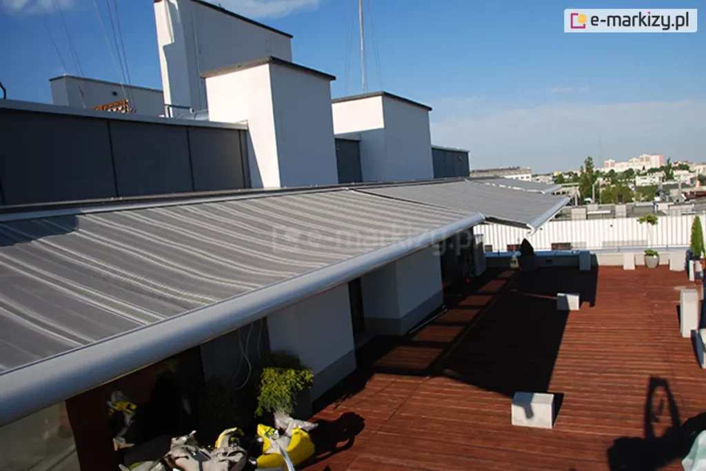 Widok z góry na rozłożony system osłonowy zainstalowany na przestronnym tarasie apartamentowca, który tworzy szereg dużych markiz Presto. Poszycie markiz wykonane jest z wysokiej jakości tkaniny w szerokie, poziome pasy w różnych odcieniach szarości, co nadaje konstrukcji nowoczesny i elegancki wygląd. Materiał jest napięty na długiej, srebrnej belce frontowej o cylindrycznym kształcie, która biegnie wzdłuż krawędzi tarasu. Markiza jest zamontowana do białych, kubikowych elementów elewacji budynku, chroniąc przed słońcem wejścia do pomieszczeń. Trzy widoczne obok siebie markizy zostały rozwinięte w różnym stopniu, by zapewnić odpowiednie zacienienie tarasu. Pod markizami znajduje się podłoga tarasu wyłożona ciemnobrązowymi deskami kompozytowymi lub drewnianymi, na których widoczne są cienie rzucane przez konstrukcję oraz betonowe donice w kształcie kostek. W tle, pod bezchmurnym błękitnym niebem, rozpościera się panorama miasta z widocznymi jasnymi budynkami mieszkalnymi i pasem zieleni w oddali.