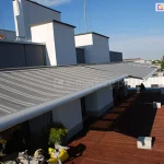 Widok z góry na rozłożony system osłonowy zainstalowany na przestronnym tarasie apartamentowca, który tworzy szereg dużych markiz Presto. Poszycie markiz wykonane jest z wysokiej jakości tkaniny w szerokie, poziome pasy w różnych odcieniach szarości, co nadaje konstrukcji nowoczesny i elegancki wygląd. Materiał jest napięty na długiej, srebrnej belce frontowej o cylindrycznym kształcie, która biegnie wzdłuż krawędzi tarasu. Markiza jest zamontowana do białych, kubikowych elementów elewacji budynku, chroniąc przed słońcem wejścia do pomieszczeń. Trzy widoczne obok siebie markizy zostały rozwinięte w różnym stopniu, by zapewnić odpowiednie zacienienie tarasu. Pod markizami znajduje się podłoga tarasu wyłożona ciemnobrązowymi deskami kompozytowymi lub drewnianymi, na których widoczne są cienie rzucane przez konstrukcję oraz betonowe donice w kształcie kostek. W tle, pod bezchmurnym błękitnym niebem, rozpościera się panorama miasta z widocznymi jasnymi budynkami mieszkalnymi i pasem zieleni w oddali.