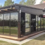 Zdjęcie przedstawia rozległą zabudowę tarasu restauracyjnego lub hotelowego, wykonaną w ciemnobrązowej konstrukcji drewnianej z dachem krytym blachodachówką. Ściany frontowe i boczne zostały w pełni przeszklone za pomocą systemu ramowego Sotro w ciemnych profilach, z dodatkowymi poprzecznymi szprosami w dolnej części skrzydeł. Jedna z sekcji szklanych jest otwarta, ukazując wnętrze z ciemnymi meblami i roślinnością. Nad częścią przeszkleń zamontowano zwinięte markizy kasetowe w kolorze bordowym. Całość posadowiona jest na drewnianym podeście z wbudowanym oświetleniem punktowym LED, otoczonym opaską z kostki brukowej i zadbanym trawnikiem.