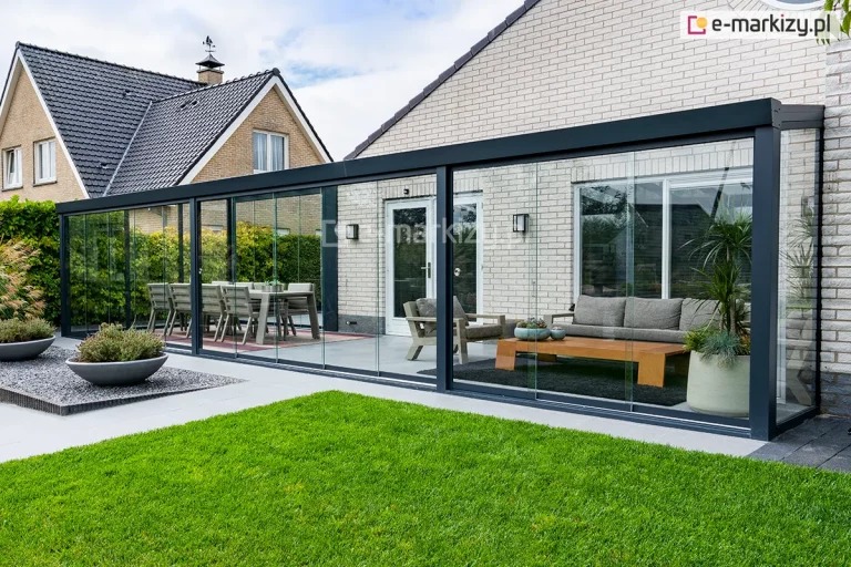 Rozbudowana szklana zabudowa tarasu w formie ogrodu letniego, wykonana na bazie aluminiowej konstrukcji Terano w kolorze antracytowym. System składa się z długiego zadaszenia szklanego opartego na ścianie budynku z jasnej cegły oraz pełnego przeszklenia bocznego i frontowego. Frontowa ściana zabudowy została wyposażona w system wieloskrzydłowych szklanych drzwi przesuwnych, które poruszają się po wielotorowych prowadnicach dolnych. Wewnątrz przestronnego wnętrza wydzielono dwie strefy: jadalnianą z dużym stołem i krzesłami oraz wypoczynkową z szarą sofą, fotelem i niskim drewnianym stolikiem kawowym ustawionym na ciemnym dywanie. Podłoże tarasu stanowią gładkie, jasnoszare płyty, a przed samą konstrukcją znajduje się zadbany trawnik oraz ozdobne donice z niską roślinnością ustawione na wysypisku z ciemnego żwiru. W tle widoczna jest architektura osiedla domów jednorodzinnych pod lekko zachmurzonym niebem.