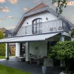 Widok z boku, pod lekkim kątem na dwie szerokie, rozciągnięte osłony przeciwsłoneczne w kolorze jasnoszarym, zainstalowana pod balkonem piętrowego domu jednorodzinnego. Materiał markiz Palladio Selt jest gładki i napięty na konstrukcji z ramionami przegubowymi, tworząc zadaszenie nad parterowym tarasem. Budynek posiada białą elewację oraz charakterystyczny, wysoki dach kryty czerwoną dachówką z lukarną, a balkon na piętrze zabezpieczony jest ozdobną, kutą barierka o wygiętym kształcie. Pod zadaszeniem znajduje się zestaw mebli ogrodowych wykonanych z szarego technorattanu, w tym sofa z poduszkami oraz niski stolik kawowy, ustawione na podłodze z dużych, ciemnoszarych płyt. Teren wokół tarasu wypełnia zielony trawnik, wysokie tuje oraz gęsty krzew o dużych liściach po prawej stronie. W tle widoczny jest fragment ogrodu z drewnianą huśtawką oraz sąsiednie domy, a wszystko to uwiecznione na tle wieczornego nieba z rozproszonymi chmurami oświetlonymi zachodzącym słońcem.