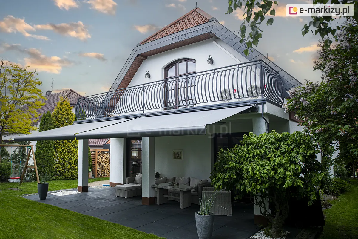 Widok z boku, pod lekkim kątem na dwie szerokie, rozciągnięte osłony przeciwsłoneczne w kolorze jasnoszarym, zainstalowana pod balkonem piętrowego domu jednorodzinnego. Materiał markiz Palladio Selt jest gładki i napięty na konstrukcji z ramionami przegubowymi, tworząc zadaszenie nad parterowym tarasem. Budynek posiada białą elewację oraz charakterystyczny, wysoki dach kryty czerwoną dachówką z lukarną, a balkon na piętrze zabezpieczony jest ozdobną, kutą barierka o wygiętym kształcie. Pod zadaszeniem znajduje się zestaw mebli ogrodowych wykonanych z szarego technorattanu, w tym sofa z poduszkami oraz niski stolik kawowy, ustawione na podłodze z dużych, ciemnoszarych płyt. Teren wokół tarasu wypełnia zielony trawnik, wysokie tuje oraz gęsty krzew o dużych liściach po prawej stronie. W tle widoczny jest fragment ogrodu z drewnianą huśtawką oraz sąsiednie domy, a wszystko to uwiecznione na tle wieczornego nieba z rozproszonymi chmurami oświetlonymi zachodzącym słońcem.