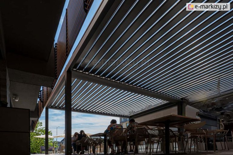 Dwie pergole lamelowe wolnostojące Sb400 Selt połączone modułowo i ustawione przy wyjściu z restauracji. Tworzą wspólnie otwierane zadaszenie przestrzeni ogródka restauracyjnego, w którym klienci spożywają posiłki na świeżym powietrzu