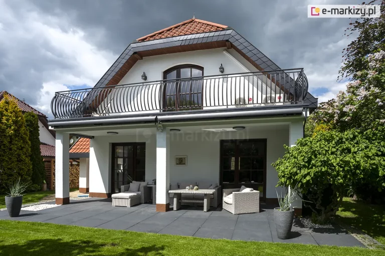 Nowoczesny budynek jednorodzinny o białej elewacji z poddaszem użytkowym i dachem wielospadowym pokrytym dachówką w kolorze ceglastym, z krawędziami wykończonymi ciemnoszarą dachówką płaską. Na piętrze znajduje się szeroki balkon z ozdobną, czarną barierką o wygiętych, pionowych prętach. Pod krawędzią balkonu zamontowane zostały dwie długie, złożone markizy tarasowe Palladio Selt w ciemnej kasecie. Parter budynku obejmuje zadaszony taras wyłożony dużymi, ciemnoszarymi płytami, na którym ustawiono jasnoszary zestaw mebli ogrodowych z technorattanu, składający się z sofy, fotela, pufy oraz stolika kawowego z jasnym blatem. Pod zadaszeniem tarasu widoczne są dwa duże przeszklone wyjścia tarasowe w ciemnych ramkach oraz dwie okrągłe lampy sufitowe. Teren wokół tarasu jest starannie zagospodarowany z zielonym trawnikiem, ozdobnymi krzewami oraz wysokimi tujami, a po bokach ustawiono dwa wysokie, szare donice z roślinami ozdobnymi. Nad budynkiem rozciąga się dynamiczne niebo z dużymi, białymi i szarymi chmurami, a po prawej stronie kadru widoczne są kwitnące krzewy bzu.