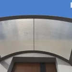 Grafika przedstawia widok z góry na łukowaty daszek naddrzwiowy, zamontowany nad wnęką wejściową nowoczesnego budynku. Konstrukcja zadaszenia składa się z ciemnobrązowej ramy aluminiowej, która dzieli powierzchnię na dwa równe, symetryczne segmenty. Wypełnienie paneli stanowi poliwęglan komorowy o dymionym, brązowym odcieniu, na którego powierzchni wyraźnie widać strukturę gęstych, równoległych żłobień biegnących wzdłuż łuku. Rama daszka posiada solidne profile brzegowe oraz pionowy szpros wzmacniający, umieszczony w centralnym punkcie konstrukcji. Zadaszenie jest przytwierdzone do jasnej, gładkiej elewacji i znacząco wystaje poza krawędź wnęki, w której osadzone są brązowe drzwi z pionowym, ciemniejszym pasem dekoracyjnym. Całość prezentowana jest na tle intensywnie błękitnego nieba z rozproszonymi, białymi chmurami. Perspektywa górna eksponuje półkolisty kształt daszka oraz system odprowadzania wody opadowej zintegrowany z bocznymi profilami ramy.