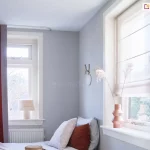 Widok na fragment jasnej sypialni utrzymanej w stonowanej, chłodnej kolorystyce z wyraźnymi akcentami w kolorze ceglanym. Na pierwszym planie po lewej stronie znajduje się wysokie okno przesłonięte gęsto marszczoną zasłoną w głębokim odcieniu rdzawym lub terakotowym, zawieszoną na cienkiej, białej szynie sufitowej. Pod oknem widoczny jest biały, żeberkowy grzejnik, a na parapecie stoi niewielka lampka nocna z jasnym, materiałowym abażurem wykończonym u dołu dekoracyjnymi frędzlami. Ściany pokoju mają kolor jasnoszary, co kontrastuje z białymi ramami okiennymi i listwami. W centralnej części obrazu, w narożniku, ustawione jest łóżko z jasną pościelą i kilkoma poduszkami, z których jedna jest w kolorze ceglanym, nawiązującym do barwy zasłon, a pozostałe są jasne i szare. Na ścianie nad łóżkiem zawieszona jest minimalistyczna dekoracja w formie okrągłego elementu z haczykiem. Po prawej stronie zdjęcia znajduje się drugie, większe okno, które zostało wyposażone w białą, półtransparentną roletę rzymską z widocznymi poziomymi fiszbinami tworzącymi regularne zakładki materiału. Na szerokim, białym parapecie tego okna ustawiono dekoracyjny wazon o geometrycznym kształcie w kolorze miedzianym, w którym umieszczono wysokie, suszone trawy ozdobne. Przez szyby wpada rozproszone, dzienne światło, a za oknami majaczą zarysy drzew i sąsiednich budynków, sugerując spokojną lokalizację. Całość wnętrza prezentuje się nowocześnie i przytulnie dzięki połączeniu chłodnych szarości z ciepłymi detalami tekstylnymi