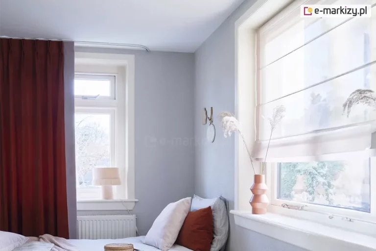 Widok na fragment jasnej sypialni utrzymanej w stonowanej, chłodnej kolorystyce z wyraźnymi akcentami w kolorze ceglanym. Na pierwszym planie po lewej stronie znajduje się wysokie okno przesłonięte gęsto marszczoną zasłoną w głębokim odcieniu rdzawym lub terakotowym, zawieszoną na cienkiej, białej szynie sufitowej. Pod oknem widoczny jest biały, żeberkowy grzejnik, a na parapecie stoi niewielka lampka nocna z jasnym, materiałowym abażurem wykończonym u dołu dekoracyjnymi frędzlami. Ściany pokoju mają kolor jasnoszary, co kontrastuje z białymi ramami okiennymi i listwami. W centralnej części obrazu, w narożniku, ustawione jest łóżko z jasną pościelą i kilkoma poduszkami, z których jedna jest w kolorze ceglanym, nawiązującym do barwy zasłon, a pozostałe są jasne i szare. Na ścianie nad łóżkiem zawieszona jest minimalistyczna dekoracja w formie okrągłego elementu z haczykiem. Po prawej stronie zdjęcia znajduje się drugie, większe okno, które zostało wyposażone w białą, półtransparentną roletę rzymską z widocznymi poziomymi fiszbinami tworzącymi regularne zakładki materiału. Na szerokim, białym parapecie tego okna ustawiono dekoracyjny wazon o geometrycznym kształcie w kolorze miedzianym, w którym umieszczono wysokie, suszone trawy ozdobne. Przez szyby wpada rozproszone, dzienne światło, a za oknami majaczą zarysy drzew i sąsiednich budynków, sugerując spokojną lokalizację. Całość wnętrza prezentuje się nowocześnie i przytulnie dzięki połączeniu chłodnych szarości z ciepłymi detalami tekstylnymi