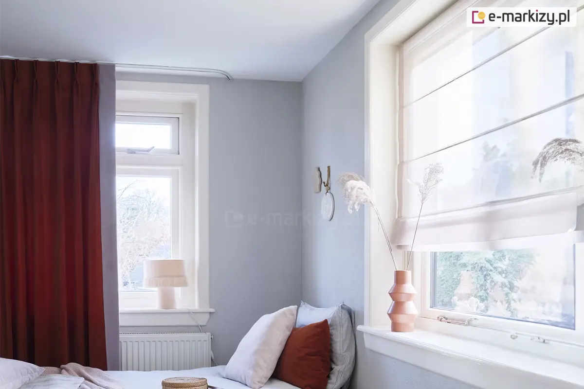 Widok na fragment jasnej sypialni utrzymanej w stonowanej, chłodnej kolorystyce z wyraźnymi akcentami w kolorze ceglanym. Na pierwszym planie po lewej stronie znajduje się wysokie okno przesłonięte gęsto marszczoną zasłoną w głębokim odcieniu rdzawym lub terakotowym, zawieszoną na cienkiej, białej szynie sufitowej. Pod oknem widoczny jest biały, żeberkowy grzejnik, a na parapecie stoi niewielka lampka nocna z jasnym, materiałowym abażurem wykończonym u dołu dekoracyjnymi frędzlami. Ściany pokoju mają kolor jasnoszary, co kontrastuje z białymi ramami okiennymi i listwami. W centralnej części obrazu, w narożniku, ustawione jest łóżko z jasną pościelą i kilkoma poduszkami, z których jedna jest w kolorze ceglanym, nawiązującym do barwy zasłon, a pozostałe są jasne i szare. Na ścianie nad łóżkiem zawieszona jest minimalistyczna dekoracja w formie okrągłego elementu z haczykiem. Po prawej stronie zdjęcia znajduje się drugie, większe okno, które zostało wyposażone w białą, półtransparentną roletę rzymską z widocznymi poziomymi fiszbinami tworzącymi regularne zakładki materiału. Na szerokim, białym parapecie tego okna ustawiono dekoracyjny wazon o geometrycznym kształcie w kolorze miedzianym, w którym umieszczono wysokie, suszone trawy ozdobne. Przez szyby wpada rozproszone, dzienne światło, a za oknami majaczą zarysy drzew i sąsiednich budynków, sugerując spokojną lokalizację. Całość wnętrza prezentuje się nowocześnie i przytulnie dzięki połączeniu chłodnych szarości z ciepłymi detalami tekstylnymi