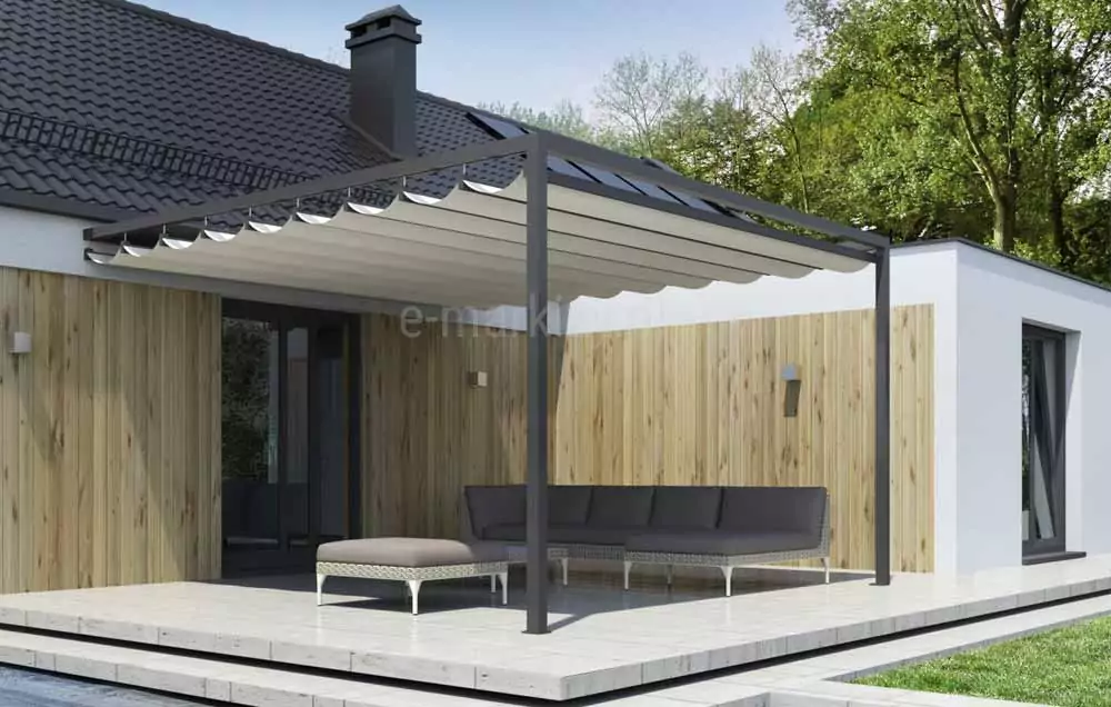 Nowoczesna pergola tarasowa Dragon przyścienna o konstrukcji aluminiowej w kolorze ciemnego antracytu zamontowana przy elewacji budynku jednorodzinnego. Zadaszenie pergoli stanowi rozsuwane poszycie z jasnego, kremowego materiału ułożonego w charakterystyczne, miękkie fałdy, które są podwieszone pod poprzecznymi profilami prowadzącymi. Konstrukcja opiera się na dwóch pionowych słupach o kwadratowym przekroju, przytwierdzonych do jasnego, kamiennego tarasu. Jedna strona pergoli jest zamontowana bezpośrednio do ściany budynku pod okapem dachu pokrytego ciemną dachówką. Ściany domu wykończone są pionowymi deskami elewacyjnymi w naturalnym, jasnym odcieniu drewna oraz białym tynkiem w bocznej części bryły. Pod zadaszeniem znajduje się zestaw mebli ogrodowych składający się z dużego, narożnego narożnika z szarymi poduszkami oraz dopasowanego pufa ze srebrną, plecioną podstawą. Taras jest wyniesiony ponad poziom ogrodu, a w tle widoczne są wysokie, zielone drzewa liściaste pod jasnym niebem.