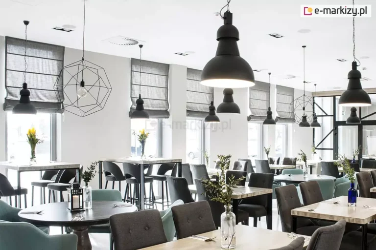Zdjęcie przedstawia przestronne i nowocześnie urządzone wnętrze restauracji lub kawiarni, w którym kluczowym elementem dekoracyjnym i funkcjonalnym są rolety rzymskie. Na długiej, białej ścianie z licznymi przeszkleniami zamontowano szereg identycznych rolet rzymskich w kolorze stonowanej, średniej szarości. Materiał rolet posiada wyraźną strukturę tkaniny, a poziome fiszbiny dzielą je na równe, estetyczne segmenty, które po podciągnięciu tworzą dekoracyjne fałdy. Rolety rzymskie są ustawione na różnych wysokościach, co pozwala na precyzyjną regulację dopływu naturalnego światła do wnętrza. Pomieszczenie jest jasne, z białym sufitem, na którym widoczne są punkty oświetleniowe oraz kratki wentylacyjne. Wyposażenie sali obejmuje różnorodne stoliki: okrągłe z ciemnymi blatami oraz prostokątne z blatami w kolorze jasnego drewna. Przy stołach ustawiono komfortowe meble tapicerowane – krzesła i fotele w kolorach ciemnoszarym oraz delikatnym błękicie, niektóre z nich ozdobione są dekoracyjnymi guzikami na oparciach. W głębi sali znajdują się również wyższe stoliki typu barowego z dopasowanymi hokerami. Oświetlenie wnętrza jest wielowarstwowe i industrialne. Z sufitu zwisają liczne czarne lampy o różnych kształtach: od dużych, solidnych kloszy w stylu loftowym, przez ażurowe, geometryczne konstrukcje z drutu, aż po proste oprawy z odsłoniętymi żarówkami. Na stolikach rozmieszczono subtelne dekoracje, takie jak szklane wazony i butelki z zielonymi gałązkami oraz żółtymi kwiatami, a także małe lampiony ze świecami. Całość kompozycji tworzy profesjonalną, a jednocześnie przytulną przestrzeń komercyjną, w której szare rolety rzymskie harmonijnie współgrają z nowoczesnym designem i chłodną paletą barw.