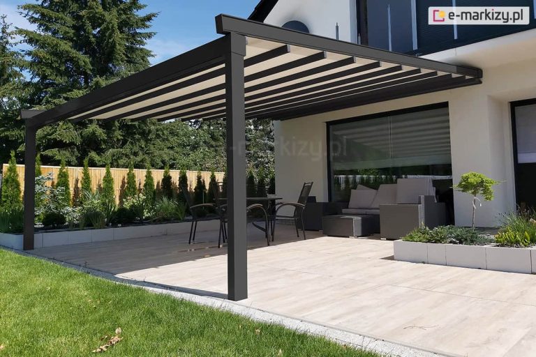 Przyścienna pergola tarasowa Square o konstrukcji aluminiowej w kolorze ciemnego antracytu, zamontowana do białej elewacji nowoczesnego budynku mieszkalnego. Zadaszenie pergoli składa się z ruchomego poszycia tkaninowego w kolorze jasnokremowym, które jest rozpięte na poprzecznych profilach, tworząc efektowne pasy. Konstrukcja wspiera się na dwóch solidnych, przednich słupach o kwadratowym przekroju, umiejscowionych na krawędzi tarasu. Pod pergolą zaaranżowano strefę wypoczynkową, w której znajduje się ciemnoszary narożnik ogrodowy z jasnymi poduszkami oraz stolik kawowy. Obok ustawiono zestaw jadalniany składający się z okrągłego stołu i metalowych krzeseł z ciemnym obiciem. Podłoże tarasu wykonane jest z dużych płyt gresowych imitujących jasne drewno. Taras z jednej strony jest otoczony niskim murkiem z białych bloczków betonowych, pełniących funkcję donic, w których posadzono niską roślinność ozdobną oraz małe drzewko formowane. W tle widoczny jest zadbany trawnik oraz gęsty szpaler tui przy drewnianym ogrodzeniu, a za nimi wysokie drzewa iglaste pod bezchmurnym, błękitnym niebem. Okno tarasowe budynku jest przesłonięte wewnętrzną roletą rzymską w kolorze dopasowanym do poszycia pergoli.