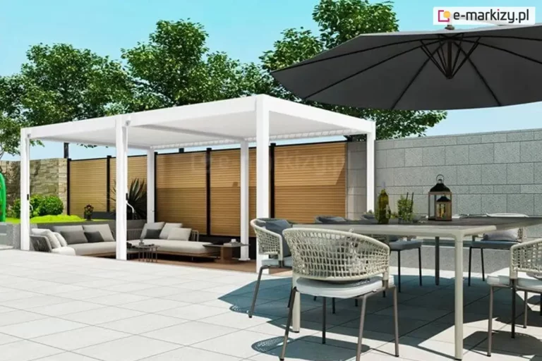 Dwumodułowa pergola wolnostojąca w kolorze białym, zainstalowana na nowoczesnym tarasie wyłożonym jasnymi płytami wielkoformatowymi. Konstrukcja o wymiarach 600 na 300 centymetrów wspiera się na sześciu słupach, a jej zadaszenie stanowią ruchome lamele aluminiowe, które w tym ujęciu są całkowicie zamknięte. Pod lewym modułem pergoli znajduje się strefa wypoczynkowa z szarą kanapą narożną ustawioną na drewnianym podeście. Na pierwszym planie, po prawej stronie, widoczna jest jadalnia pod gołym niebem z szarym parasolem bocznym, dużym stołem i krzesłami o ażurowym splocie. Architektura otoczenia obejmuje minimalistyczny budynek z dużymi przeszkleniami, szklanymi balustradami oraz zewnętrznymi schodami prowadzącymi na wyższy poziom. Całość dopełnia zadbana roślinność w postaci równo przystrzyżonego trawnika oraz formowanych w kule krzewów wzdłuż ścieżki.