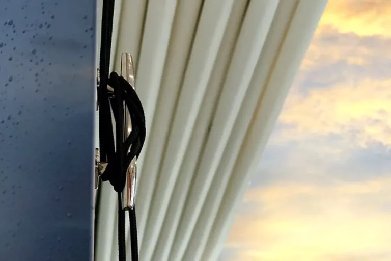 Zbliżenie stylowego elementu do zaczepiania sterowania sznurkowego dachem w pergoli Dragon. Błyszczący element został przykręcony do grafitowej podpory i opleciony czarnym sznurkiem. W tle widoczna boczna zasłona tkaninowa oraz niebo z chmurami podświetlonymi wczesnoporannym, żółtym blaskiem słońca.