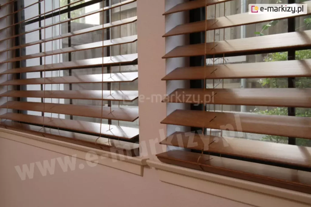 Zdjęcie prezentuje perspektywiczny widok na dwa sąsiadujące okna, w których zamontowane są żaluzje drewniane w kolorze średniego, ciepłego brązu. Kadrowanie z niskiego kąta pozwala na szczegółowe zaobserwowanie układu lameli, które są ustawione w pozycji lekko uchylonej, co tworzy rytmiczny wzór nachylonych płaszczyzn. Światło padające z zewnątrz odbija się od gładkiej, satynowej powierzchni drewna, uwypuklając jego naturalną strukturę. Przez szczeliny między listwami widoczne jest tło za oknem, obejmujące fragmenty drewnianego ogrodzenia oraz zielone liście roślin ogrodowych. Przez lamele przebiegają pionowe, cienkie drabinki sznurkowe w kolorze brązowym, które stabilizują konstrukcję. Okna posiadają jasne ramy i są osadzone w ścianie o delikatnym, brzoskwiniowym lub beżowym odcieniu, a na dole wykończone są klasycznymi, białymi parapetami. Całość kompozycji podkreśla, jak żaluzje drewniane skutecznie regulują dopływ światła, jednocześnie stanowiąc elegancki i naturalny element wykończenia wnętrza.