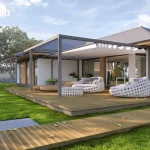 Wizualizacja przedstawia nowoczesną, rozbudowaną pergolę tarasową Tarasola Flat o konstrukcji modułowej, zamontowaną na dużym tarasie z naturalnych desek drewnianych. Ciemnoszara, aluminiowa rama o matowym wykończeniu składa się z dwóch sekcji, na których tkanina zadaszenia pozostaje częściowo odsłonięta, ukazując precyzyjny system prowadnic. Pod zadaszoną częścią zaaranżowano luksusową strefę wypoczynku z dwoma dużymi, okrągłymi fotelami ogrodowymi o ażurowej, białej konstrukcji oraz dopasowanym stolikiem kawowym. W głębi widoczne są również drewniane leżaki tarasowe. Pergola przylega do parterowego budynku o białej elewacji z drewnianymi akcentami i szerokimi przeszkleniami, które zacierają granicę między wnętrzem a ogrodem. Otoczenie stanowi rozległy, intensywnie zielony trawnik z widocznym w oddali murkiem gabionowym oraz pasem gęstych drzew, co nadaje całości kompozycji prestiżowy i spokojny charakter.