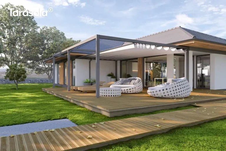 Wizualizacja przedstawia nowoczesną, rozbudowaną pergolę tarasową Tarasola Flat o konstrukcji modułowej, zamontowaną na dużym tarasie z naturalnych desek drewnianych. Ciemnoszara, aluminiowa rama o matowym wykończeniu składa się z dwóch sekcji, na których tkanina zadaszenia pozostaje częściowo odsłonięta, ukazując precyzyjny system prowadnic. Pod zadaszoną częścią zaaranżowano luksusową strefę wypoczynku z dwoma dużymi, okrągłymi fotelami ogrodowymi o ażurowej, białej konstrukcji oraz dopasowanym stolikiem kawowym. W głębi widoczne są również drewniane leżaki tarasowe. Pergola przylega do parterowego budynku o białej elewacji z drewnianymi akcentami i szerokimi przeszkleniami, które zacierają granicę między wnętrzem a ogrodem. Otoczenie stanowi rozległy, intensywnie zielony trawnik z widocznym w oddali murkiem gabionowym oraz pasem gęstych drzew, co nadaje całości kompozycji prestiżowy i spokojny charakter.