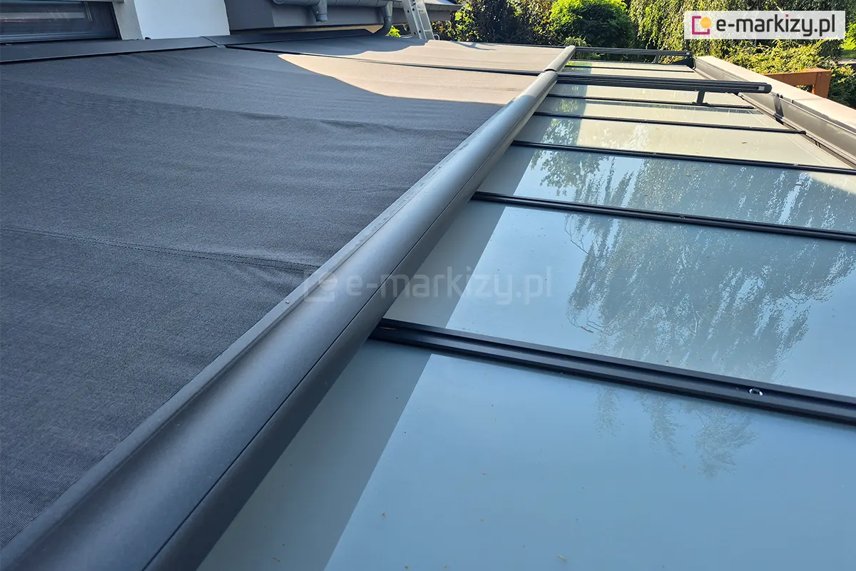 Perspektywiczne ujęcie systemu Veranda składającego się z dwóch modułów, zamontowanego nad szklanym zadaszeniem tarasu. Na zdjęciu widoczna jest antracytowa, aluminiowa belka przednia (obciążająca), która wysuwa ciemnoszarą tkaninę techniczną wzdłuż czarnych prowadnic bocznych. Markiza jest w połowie rozwinięta, odsłaniając tafle szkła, w których odbijają się korony drzew i niebo. Konstrukcja prowadnic jest uniesiona nad powierzchnią szkła za pomocą systemowych wsporników. W tle widoczny jest fragment elewacji budynku, rynna spustowa oraz gęsta zieleń ogrodowa. Zdjęcie ilustruje płynne działanie mechanizmu oraz precyzyjne napięcie materiału osłonowego.