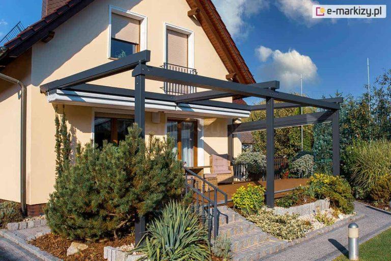 Nowoczesna pergola tarasowa przyścienna o konstrukcji aluminiowej w kolorze antracytowym, przedstawiona ze zwiniętym, jasnym poszyciem tkaninowym, zamontowana przy beżowej elewacji domu jednorodzinnego. Aluminiowy stelaż opiera się na solidnych profilach pionowych, a zadaszenie segmentowe wsparte jest na poprzecznych belkach, tworząc zintegrowaną osłonę nad tarasem wykonanym z brązowych desek kompozytowych. Na taras prowadzą kamienne schody z szarego granitu wyposażone w metalową balustradę, a w strefie wypoczynkowej pod pergolą widoczne są plecione fotele ogrodowe z jasnymi poduszkami. Wokół tarasu znajduje się bogato zaaranżowany ogród z dużą sosną górską, juką oraz niskimi krzewami ozdobnymi posadzonymi na podłożu z kory sosnowej, oddzielonym od ścieżki z kostki brukowej kamiennymi krawężnikami. Wzdłuż traktu komunikacyjnego ustawiono nowoczesne lampy ogrodowe w formie srebrnych słupków, a w tle widoczne są okna budynku z opuszczonymi roletami zewnętrznymi oraz błękitne niebo z delikatnymi chmurami.