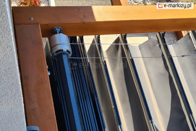 Elektryczny dach rzymski Supro do konstrukcji drewnianej pergola koro z widocznym elementem napędu oraz linkami stalowymi oraz układającą się w fale tkaniną Opatex w kolorze antracytowym.