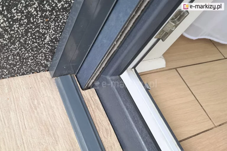 Płaski próg PCV moskitiery Click Rol zamocowany do płytki tarasowej w drewnopodobnym kolorze w pobliżu ramy okna i prowadnic rolety zewnętrznej. Wszystkie elementy wykonane w odcieniach antracytu.