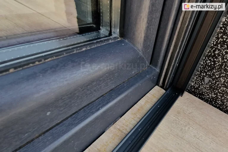 Drewniana rama okna o antracytowym kolorze, prowadnica rolety zewnętrznej z widocznymi uszczelkami szczotkowymi oraz profil boczny moskitiery plisowanej Top Zag zamocowanej do wnęki o szarej elewacji. Moskitiera wyposażona w niski próg z ukrytymi w nim sznurkami, naklejony na płytki gresowe o drewnianym wzorze ułożone na tarasie przed oknem.