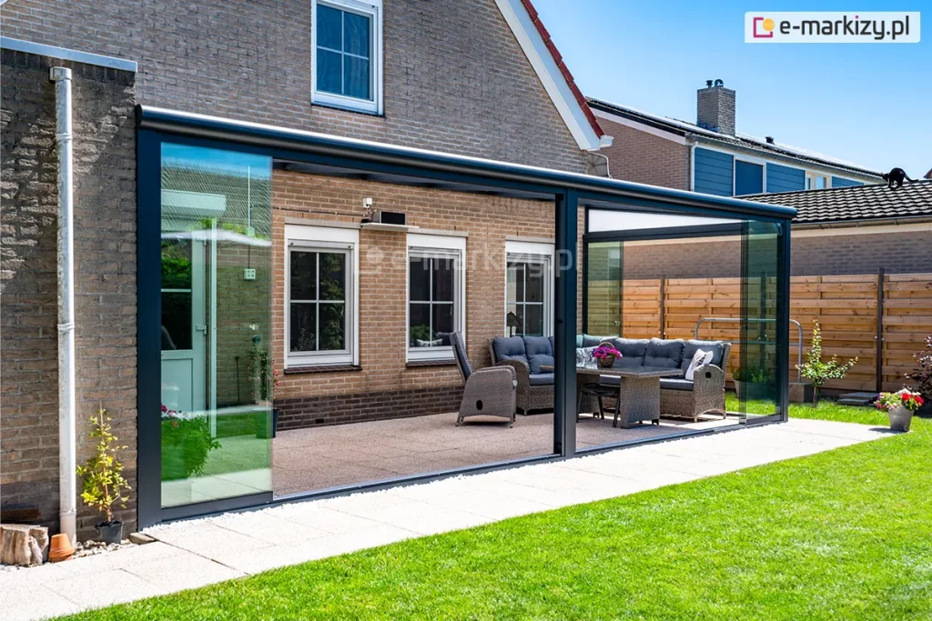 Zdjęcie prezentuje zewnętrzne ujęcie nowoczesnego ogrodu letniego opartego na antracytowej konstrukcji zadaszenia tarasowego Bosco, zamontowanego przy domu z jasnobrązowej cegły. Dach konstrukcji składa się z aluminiowych krokwi z wypełnieniem z mlecznego, półprzezroczystego poliwęglanu, a front i boki zostały zabudowane systemem bezramowych szklanych ścian przesuwnych Auris, które chronią wnętrze przed wiatrem i deszczem. Wewnątrz przeszklonej przestrzeni znajduje się strefa wypoczynkowa z dużą sofą z szarego technorattanu, dopasowanym fotelem oraz stołem z doniczką różowych kwiatów, ustawionymi na tarasie wyłożonym płytkami o drobnym wzorze. Panele szklane na obu połowach frontowych przesłon zostały odsunięte, co otwiera taras na zadbany ogród z zielonym trawnikiem i drewnianym ogrodzeniem w tle. Całość dopełnia widok na sąsiednie budynki pod bezchmurnym, błękitnym niebem oraz detale elewacji domu z białymi ramami okiennymi.