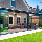 Zdjęcie prezentuje zewnętrzne ujęcie nowoczesnego ogrodu letniego opartego na antracytowej konstrukcji zadaszenia tarasowego Bosco, zamontowanego przy domu z jasnobrązowej cegły. Dach konstrukcji składa się z aluminiowych krokwi z wypełnieniem z mlecznego, półprzezroczystego poliwęglanu, a front i boki zostały zabudowane systemem bezramowych szklanych ścian przesuwnych Auris, które chronią wnętrze przed wiatrem i deszczem. Wewnątrz przeszklonej przestrzeni znajduje się strefa wypoczynkowa z dużą sofą z szarego technorattanu, dopasowanym fotelem oraz stołem z doniczką różowych kwiatów, ustawionymi na tarasie wyłożonym płytkami o drobnym wzorze. Panele szklane na obu połowach frontowych przesłon zostały odsunięte, co otwiera taras na zadbany ogród z zielonym trawnikiem i drewnianym ogrodzeniem w tle. Całość dopełnia widok na sąsiednie budynki pod bezchmurnym, błękitnym niebem oraz detale elewacji domu z białymi ramami okiennymi.