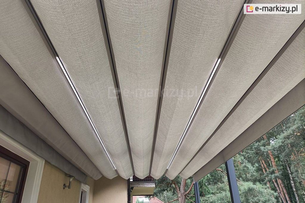 Zbliżenie dachu pergoli Tako z oświetleniem led w belkach przeciwwietrznych oraz akrylową tkaniną poszycia o splocie grubego płótna w kolorze jasnego beżu. Pod pergola widoczny fragment elewacji po lewej z wnękami okiennymi i lampką między nimi. W tle widoczna płyta balkonu na piętrze. Po prawej pod pergolą widoczne drzewa iglaste.