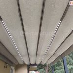 Zbliżenie dachu pergoli Tako z oświetleniem led w belkach przeciwwietrznych oraz akrylową tkaniną poszycia o splocie grubego płótna w kolorze jasnego beżu. Pod pergola widoczny fragment elewacji po lewej z wnękami okiennymi i lampką między nimi. W tle widoczna płyta balkonu na piętrze. Po prawej pod pergolą widoczne drzewa iglaste.