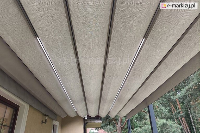 Zbliżenie dachu pergoli Tako z oświetleniem led w belkach przeciwwietrznych oraz akrylową tkaniną poszycia o splocie grubego płótna w kolorze jasnego beżu. Pod pergola widoczny fragment elewacji po lewej z wnękami okiennymi i lampką między nimi. W tle widoczna płyta balkonu na piętrze. Po prawej pod pergolą widoczne drzewa iglaste.