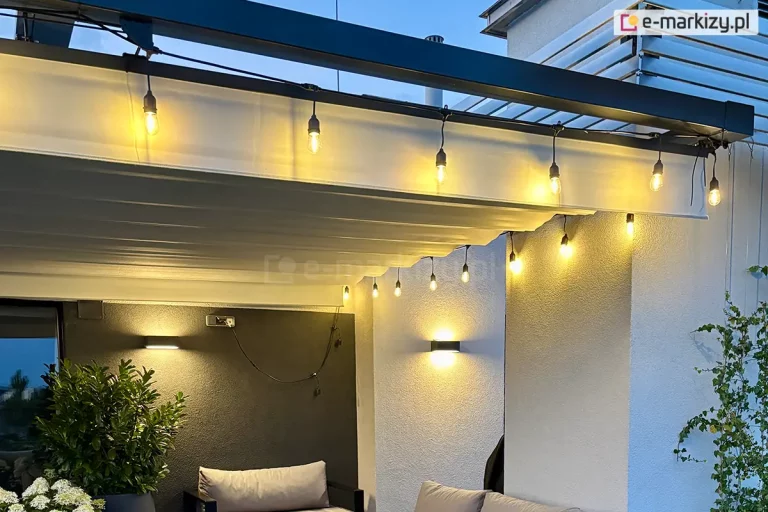 zbliżenie na system zadaszenia tarasu w formie dachu rzymskiego Supro, zamontowanego pod czarną, metalową belką konstrukcyjną. Jasnokremowa tkanina osłony jest rozłożona poziomo, tworząc charakterystyczne, miękkie pofałdowania. Wzdłuż krawędzi zadaszenia poprowadzono girlandę świetlną z żarówkami typu Edison, które emitują ciepłe, jasne światło, oświetlając materiał oraz przestrzeń poniżej. Na jasnej elewacji budynku widoczne są dwa nowoczesne, czarne kinkiety ścienne, które świecą góra-dół, podkreślając strukturę tynku. W dolnej części kadru widoczne są fragmenty mebli ogrodowych – szare poduszki sofy oraz ciemna donica z gęstą, zieloną roślinnością i białymi kwiatami (hortensje). Po prawej stronie pnie się po ścianie zielone pnącze.