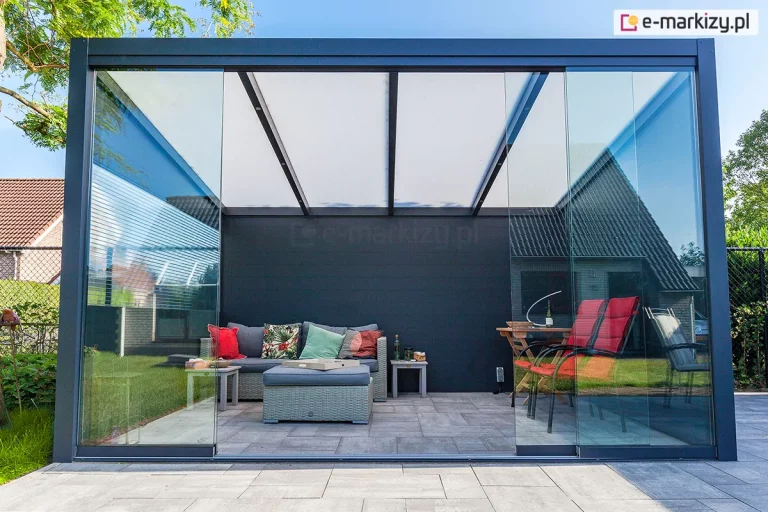 Nowoczesna zabudowa tarasu o konstrukcji aluminiowej w kolorze antracytowym. Zdjęcie prezentuje front patio z zadaszeniem z mlecznego poliwęglanu oraz systemem bezramowych szklanych drzwi przesuwnych Auris . Skrzydła drzwiowe są rozsuniete na boki, otwierając wnętrze na ogród. Wewnątrz przeszklonej altany znajduje się strefa wypoczynkowa z narożnikiem z szarego technorattanu, ozdobionymi kolorowymi poduszkami oraz stolikami pomocniczymi. Po prawej stronie widoczne są dwa fotele z czerwonymi siedziskami przy drewnianym stoliku. Podłoże wykonane jest z dużych, jasnoszarych płyt betonowych, a w tle widoczna jest czarna ściana budynku oraz zieleń ogrodowa.