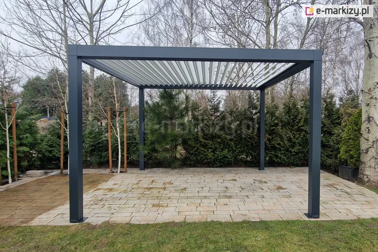 Nowoczesna pergola tarasowa wolnostojąca SB350 Selt o konstrukcji aluminiowej w kolorze ciemnoszarym (antracytowym), zamontowana na podłożu z jasnych płyt betonowych. Zadaszenie pergoli stanowią ruchome lamele w kolorze białym, ustawione pod kątem, co pozwala na regulację dopływu światła słonecznego. Konstrukcja opiera się na czterech solidnych, kwadratowych słupach przytwierdzonych do podłoża za pomocą stóp montażowych. W tle widoczny jest ogród z gęstą roślinnością, obejmującą iglaki, krzewy oraz wysokie, bezlistne drzewa liściaste w okresie wczesnowiosennym lub jesiennym. Po prawej stronie zdjęcia znajduje się duża donica z rośliną, a po lewej młode drzewka zabezpieczone tyczkami bambusowymi.