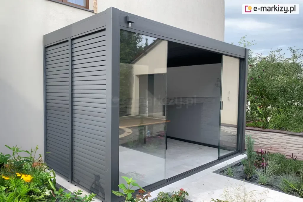 Zdjęcie przedstawia nowoczesną, wolnostojącą pergolę tarasową o konstrukcji aluminiowej w kolorze ciemnego grafitu lub antracytu, która została zamontowana bezpośrednio przy jasnej elewacji budynku. Konstrukcja charakteryzuje się minimalistycznym, kubistycznym kształtem z wyraźnie zaznaczonymi szerokimi profilami poziomymi i pionowymi. Lewa ściana boczna pergoli została wypełniona stałymi panelami z poziomych lameli aluminiowych, które pełnią funkcję przesłony przeciwsłonecznej oraz zapewniają prywatność. Frontowa część pergoli jest wyposażona w system przesuwnych przeszkleń bezramowych, które pozwalają na całkowite zamknięcie lub otwarcie przestrzeni. Na zdjęciu szklane panele są częściowo rozsunięte, ukazując wnętrze wyłożone jasnoszarymi płytami tarasowymi. Wewnątrz konstrukcji widoczny jest fragment drewnianego stołu oraz gładka, biała ściana budynku stanowiąca tylną część zabudowy. Całość posadowiona jest na przygotowanym podłożu z jasnego betonu lub kamienia, a wokół pergoli znajduje się zadbana roślinność ogrodowa, w tym zielone krzewy, żółte kwiaty oraz ozdobne trawy posadzone w pasie żwiru wzdłuż krawędzi tarasu. Tło stanowi fragment sąsiedniego budynku o jasnej elewacji oraz lekko zachmurzone niebo.