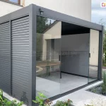 Zdjęcie przedstawia nowoczesną, wolnostojącą pergolę tarasową o konstrukcji aluminiowej w kolorze ciemnego grafitu lub antracytu, która została zamontowana bezpośrednio przy jasnej elewacji budynku. Konstrukcja charakteryzuje się minimalistycznym, kubistycznym kształtem z wyraźnie zaznaczonymi szerokimi profilami poziomymi i pionowymi. Lewa ściana boczna pergoli została wypełniona stałymi panelami z poziomych lameli aluminiowych, które pełnią funkcję przesłony przeciwsłonecznej oraz zapewniają prywatność. Frontowa część pergoli jest wyposażona w system przesuwnych przeszkleń bezramowych, które pozwalają na całkowite zamknięcie lub otwarcie przestrzeni. Na zdjęciu szklane panele są częściowo rozsunięte, ukazując wnętrze wyłożone jasnoszarymi płytami tarasowymi. Wewnątrz konstrukcji widoczny jest fragment drewnianego stołu oraz gładka, biała ściana budynku stanowiąca tylną część zabudowy. Całość posadowiona jest na przygotowanym podłożu z jasnego betonu lub kamienia, a wokół pergoli znajduje się zadbana roślinność ogrodowa, w tym zielone krzewy, żółte kwiaty oraz ozdobne trawy posadzone w pasie żwiru wzdłuż krawędzi tarasu. Tło stanowi fragment sąsiedniego budynku o jasnej elewacji oraz lekko zachmurzone niebo.