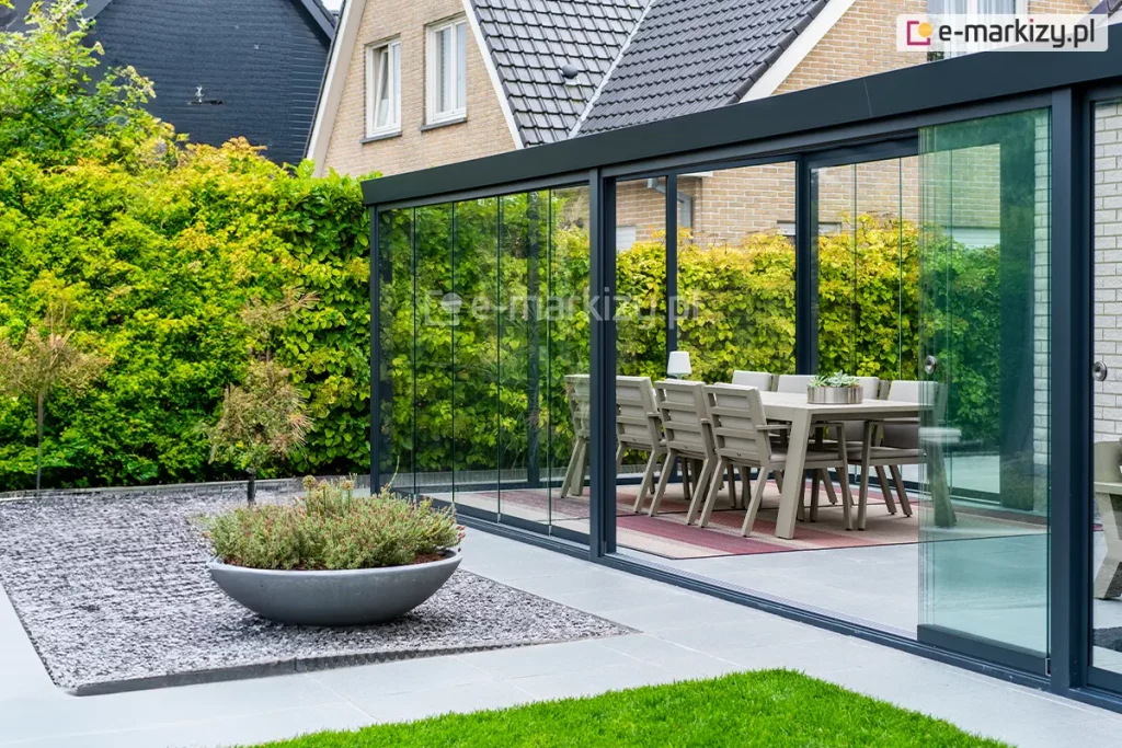 Szerokie zadaszenie tarasu w systemie przesuwnym, tworzące w pełni przeszklony ogród letni. Konstrukcja o profilach aluminiowych w kolorze antracytowym jest zamontowana do elewacji budynku wykonanej z jasnej cegły. Frontowa ściana zabudowy składa się z wieloskrzydłowych, bezramowych drzwi szklanych, które poruszają się po dolnych szynach jezdnych. Jedno ze skrzydeł jest przesunięte, ukazując sposób nakładania się tafli szkła na siebie. Na szklanych panelach zamontowano dyskretne, okrągłe uchwyty ze stali nierdzewnej. Wewnątrz ogrodu letniego znajduje się strefa jadalniana z dużym stołem i nowoczesnymi krzesłami ogrodowymi w kolorze beżowym, ustawionymi na pasiastym dywanie zewnętrznym. Podłoga tarasu wyłożona jest gładkimi płytami betonowymi w kolorze jasnoszarym. Na zewnątrz, przed konstrukcją, widoczna jest niska roślinność w ozdobnej, półkolistej misie ustawionej na polu z drobnego, ciemnego tłucznia. Pomiędzy gazonem z misą i ścieżkami widoczne zadbane zielone trawniki. W tle widać gęsty, wysoki żywopłot oraz fragmenty dachów sąsiednich budynków o ciemnym i jasnym wybarwieniu.