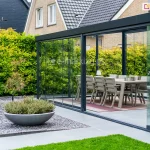 Szerokie zadaszenie tarasu w systemie przesuwnym, tworzące w pełni przeszklony ogród letni. Konstrukcja o profilach aluminiowych w kolorze antracytowym jest zamontowana do elewacji budynku wykonanej z jasnej cegły. Frontowa ściana zabudowy składa się z wieloskrzydłowych, bezramowych drzwi szklanych, które poruszają się po dolnych szynach jezdnych. Jedno ze skrzydeł jest przesunięte, ukazując sposób nakładania się tafli szkła na siebie. Na szklanych panelach zamontowano dyskretne, okrągłe uchwyty ze stali nierdzewnej. Wewnątrz ogrodu letniego znajduje się strefa jadalniana z dużym stołem i nowoczesnymi krzesłami ogrodowymi w kolorze beżowym, ustawionymi na pasiastym dywanie zewnętrznym. Podłoga tarasu wyłożona jest gładkimi płytami betonowymi w kolorze jasnoszarym. Na zewnątrz, przed konstrukcją, widoczna jest niska roślinność w ozdobnej, półkolistej misie ustawionej na polu z drobnego, ciemnego tłucznia. Pomiędzy gazonem z misą i ścieżkami widoczne zadbane zielone trawniki. W tle widać gęsty, wysoki żywopłot oraz fragmenty dachów sąsiednich budynków o ciemnym i jasnym wybarwieniu.