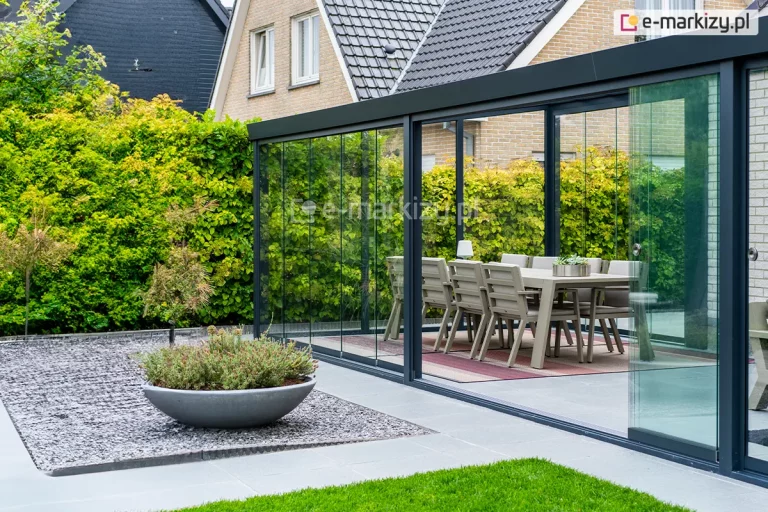 Szerokie zadaszenie tarasu w systemie przesuwnym, tworzące w pełni przeszklony ogród letni. Konstrukcja o profilach aluminiowych w kolorze antracytowym jest zamontowana do elewacji budynku wykonanej z jasnej cegły. Frontowa ściana zabudowy składa się z wieloskrzydłowych, bezramowych drzwi szklanych, które poruszają się po dolnych szynach jezdnych. Jedno ze skrzydeł jest przesunięte, ukazując sposób nakładania się tafli szkła na siebie. Na szklanych panelach zamontowano dyskretne, okrągłe uchwyty ze stali nierdzewnej. Wewnątrz ogrodu letniego znajduje się strefa jadalniana z dużym stołem i nowoczesnymi krzesłami ogrodowymi w kolorze beżowym, ustawionymi na pasiastym dywanie zewnętrznym. Podłoga tarasu wyłożona jest gładkimi płytami betonowymi w kolorze jasnoszarym. Na zewnątrz, przed konstrukcją, widoczna jest niska roślinność w ozdobnej, półkolistej misie ustawionej na polu z drobnego, ciemnego tłucznia. Pomiędzy gazonem z misą i ścieżkami widoczne zadbane zielone trawniki. W tle widać gęsty, wysoki żywopłot oraz fragmenty dachów sąsiednich budynków o ciemnym i jasnym wybarwieniu.