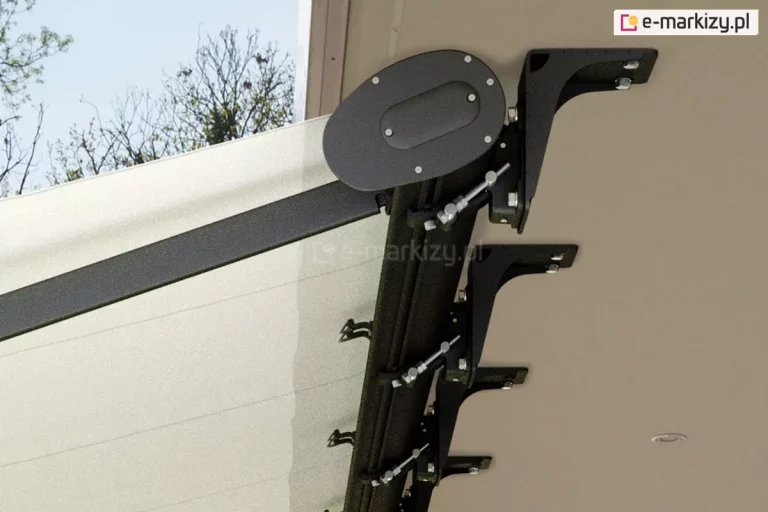 Zdjęcie prezentuje techniczne zbliżenie na system mocowania markizy Casablanca Aluprof, ukazane z perspektywy dolnej. Widoczny jest mechanizm montażowy przytwierdzony do spodu betonowego zadaszenia lub podbitki, składający się z serii solidnych, czarnych wsporników wykonanych z aluminium o porowatej strukturze. Każdy wspornik jest przymocowany do sufitu za pomocą stalowych śrub, a do nich za pomocą specjalnych klamer i śrub regulacyjnych przytwierdzona jest kaseta markizy. Po prawej stronie kadru widoczna jest boczna pokrywa kasety markizy Casablanca o owalnym kształcie, wykończona dyskretnymi nitami lub śrubami, również w kolorze matowego antracytu. Od konstrukcji odchodzi profilowane ramię przegubowe, które podtrzymuje jasne, białe poszycie tkaniny. Materiał markizy jest czysty i precyzyjnie napięty, a jego krawędź chowa się wewnątrz profilu kasety. W tle, po lewej stronie, widać fragmenty bezlistnych koron drzew na tle jasnego nieba, co sugeruje wczesnowiosenny lub jesienny okres montażu. Zdjęcie ma charakter instruktażowy, szczegółowo pokazując rozmieszczenie punktów podparcia i stabilność całej konstrukcji markizy Casablanca.