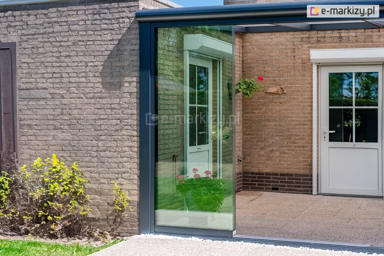 Nowoczesna zabudowa ganku lub tarasu z wykorzystaniem systemu szklanych drzwi przesuwnych Auris. Głównym elementem są wielkoformatowe, bezramowe panele ze szkła hartowanego, które są osadzone w dolnej prowadnicy zamontowanej na poziomie podłoża oraz w górnej szynie zintegrowanej z konstrukcją zadaszenia. Profile aluminiowe systemu, w tym pionowy słupek przyścienny i rynna, mają matowe wykończenie w kolorze ciemnego antracytu. W tle widoczna jest elewacja budynku wykonana z jasnobrązowej cegły oraz białe drzwi wejściowe z przeszkleniami i szprosami, wyposażone w rolety zewnętrzne. Posadzka ganku jest wyłożona jasnymi płytami tarasowymi, a przed szklaną ścianą znajduje się pas dekoracyjnego białego żwiru oraz fragment trawnika z niską roślinnością. Całość tworzy estetyczną, transparentną barierę chroniącą przed czynnikami atmosferycznymi, nie ograniczając widoku na otoczenie.