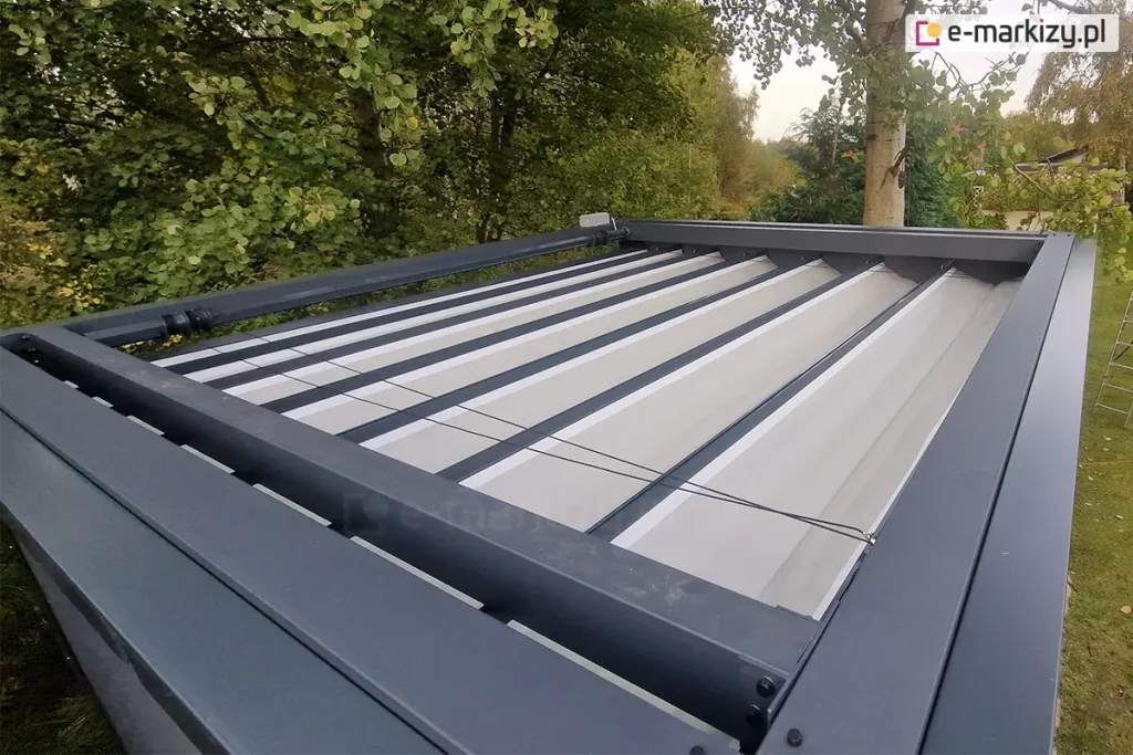 Górna powierzchnia pergoli Tako wolnostojącej. Widoczna antracytowa konstrukcja oraz prowadnice dachu,w którym zamocowane są poprzeczne belki przeciwwietrzne z fałdami jasnej tkaniny dachu. Pomiędzy elementami konstrukcji widoczne sznurki zaczepione o rurę napędową z silnikiem. Dookoła pergoli liczne drzewa i krzewy.