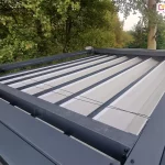 Górna powierzchnia pergoli Tako wolnostojącej. Widoczna antracytowa konstrukcja oraz prowadnice dachu,w którym zamocowane są poprzeczne belki przeciwwietrzne z fałdami jasnej tkaniny dachu. Pomiędzy elementami konstrukcji widoczne sznurki zaczepione o rurę napędową z silnikiem. Dookoła pergoli liczne drzewa i krzewy.