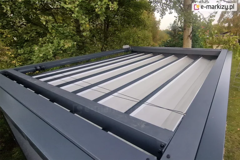 Górna powierzchnia pergoli Tako wolnostojącej. Widoczna antracytowa konstrukcja oraz prowadnice dachu,w którym zamocowane są poprzeczne belki przeciwwietrzne z fałdami jasnej tkaniny dachu. Pomiędzy elementami konstrukcji widoczne sznurki zaczepione o rurę napędową z silnikiem. Dookoła pergoli liczne drzewa i krzewy.